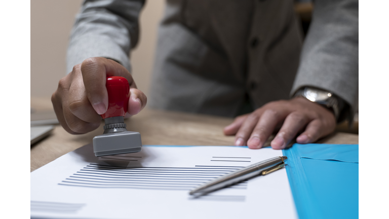 Close-up Of A Person's Hand Stamping With Approved Stamp On Document At Desk