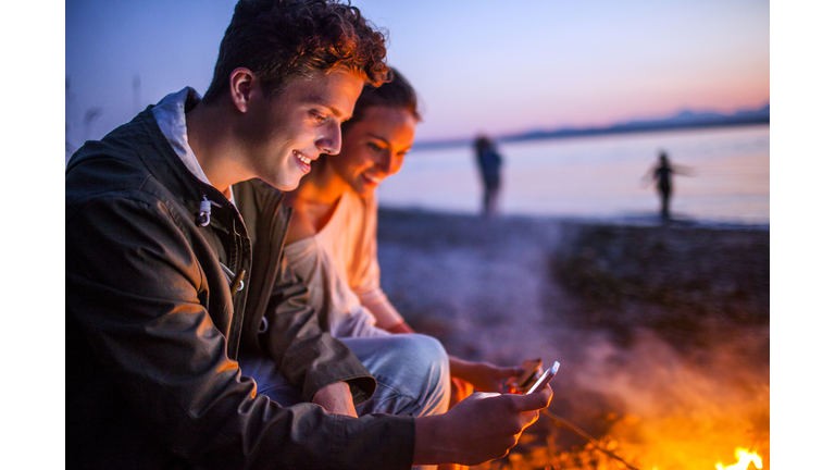 Caucasian couple using cell phone together near fire on beach