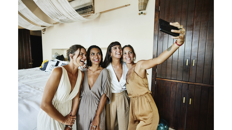 Medium wide shot of smiling and laughing bridesmaids taking selfie in luxury hotel suite before wedding