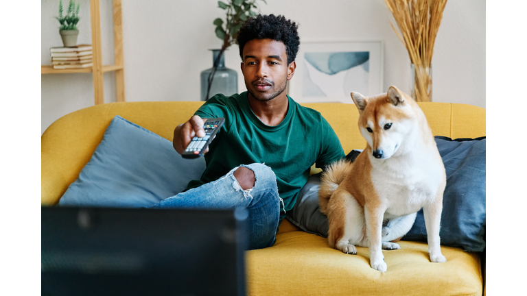 Man sitting on sofa in living room with dog watching TV changing channels with remote control.