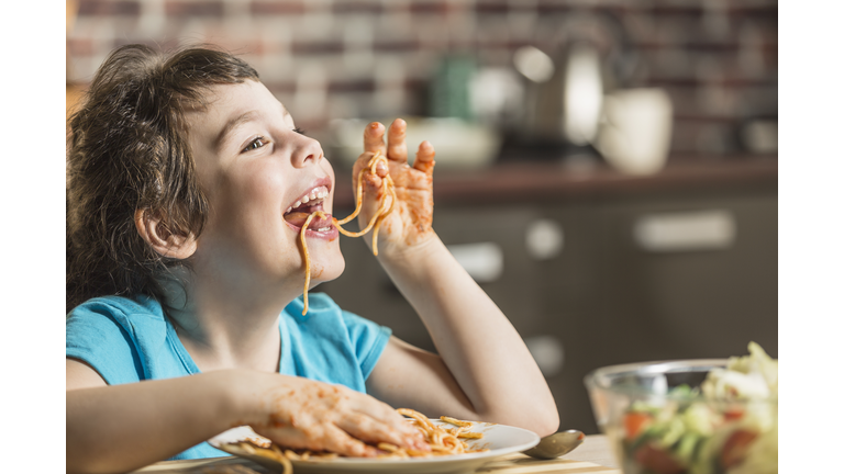 Cheerful little girl eating spaghetti with hands at home