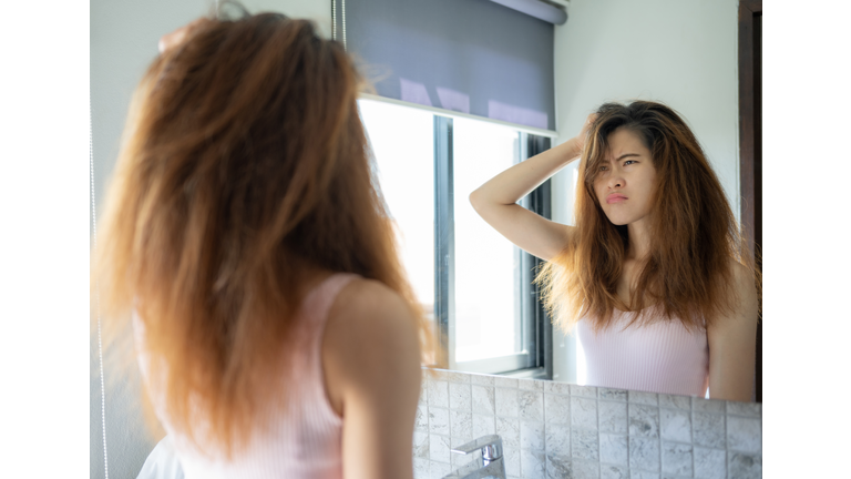 Young worried Asian woman while she saw her messy hair in the mirror after waking up in the morning.