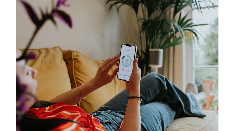 Relaxed woman reclines on a sofa and uses a thermostat app on her smart phone to control the housing heating system