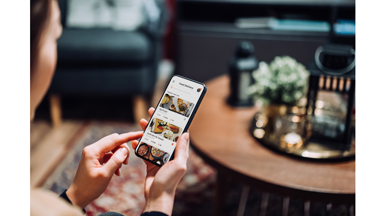 Close up of young Asian woman using meal delivery service and ordering food online with mobile app on smartphone while sitting on the sofa in the living room at a cozy home