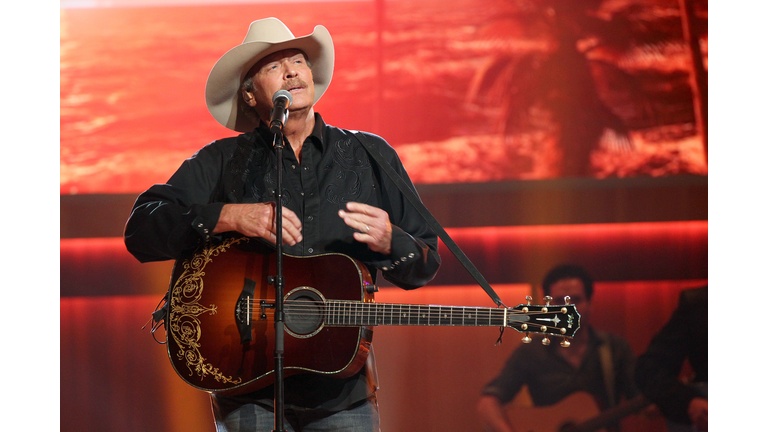 Alan Jackson in a cream colored cowboy hat, black shirt, and jeans against a sunset background.