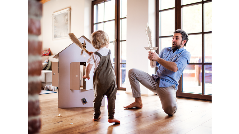 A toddler boy and father with carton swords playing indoors at home.