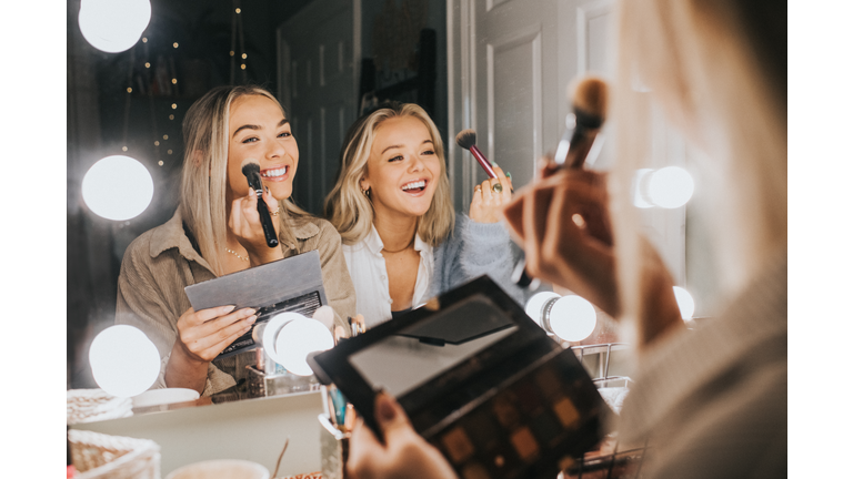 Two young woman sit in front of an illuminated mirror and apply make-up