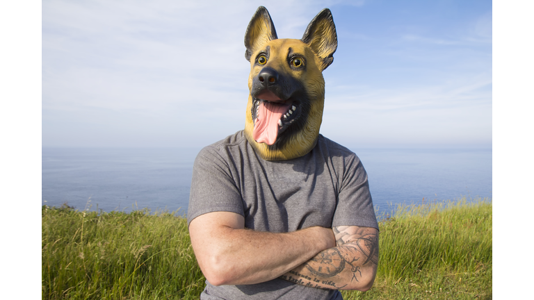 Man with tattooed arm and German Shepherd dog mask with arms crossed by the sea