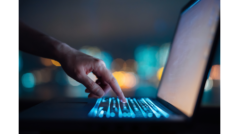 Close up of woman's hand typing on computer keyboard in the dark against colourful bokeh in background, working late on laptop at home