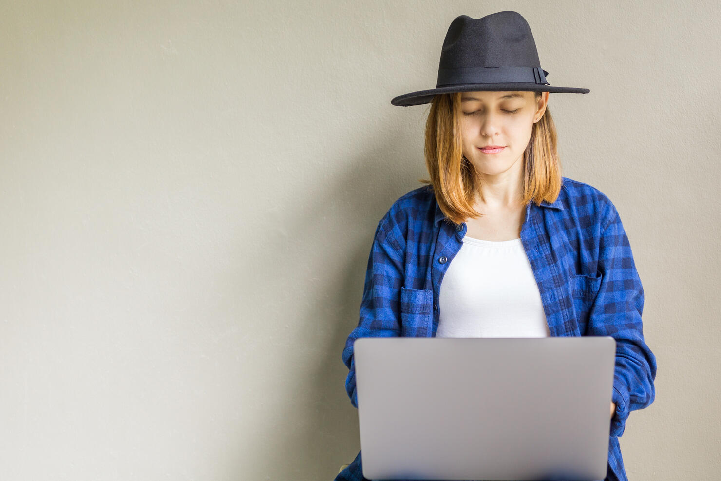Happy hipster young female wear a hat sitting in the floor working on laptop. Portrait of creative young smiling woman using computer notebook. Beautiful girl sitting on the floor near gray wall.