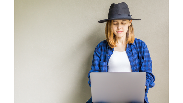 Happy hipster young female wear a hat sitting in the floor working on laptop. Portrait of creative young smiling woman using computer notebook. Beautiful girl sitting on the floor near gray wall.