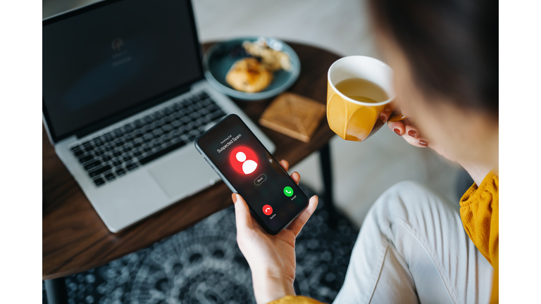 Over the shoulder view of young Asian woman receiving an incoming suspected call from unknown caller on her smartphone and rejecting the call at home. Device screen showing warning sign as detected by the network provider. Phone scam and fraud concept