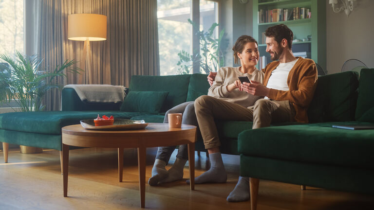 Couple Use Smartphone Device, while Sitting on a Couch in the Cozy Apartment. Boyfriend and Girlfriend Talk, do e-Shopping on Internet, Watching Funny Videos, Use Social Media, Streaming Service.