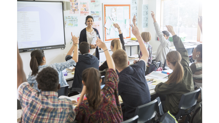 Rear view of teenage students raising hands in classroom