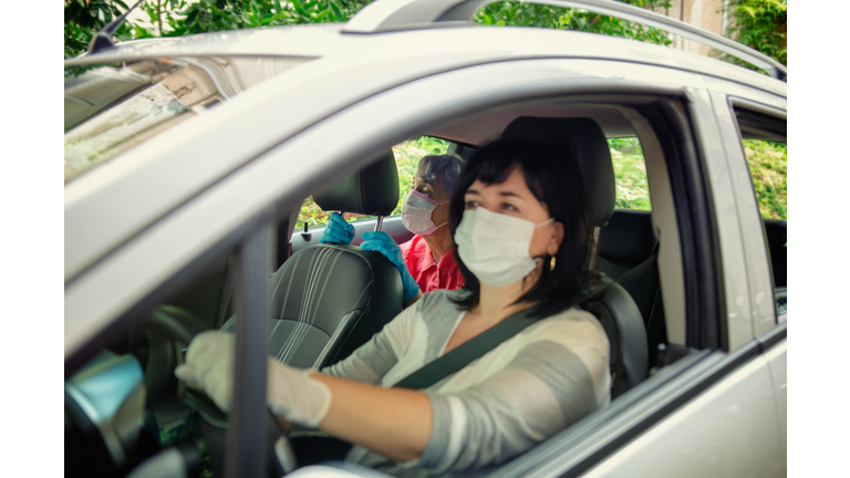 Caregiver drives a senior adult woman in a white car for daily medical procedures. Both put on protective masks.