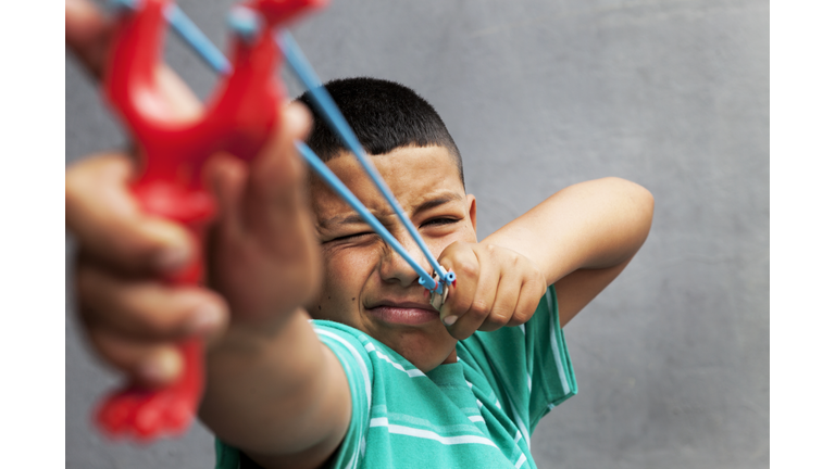 Hispanic kid pointing with a slingshot.