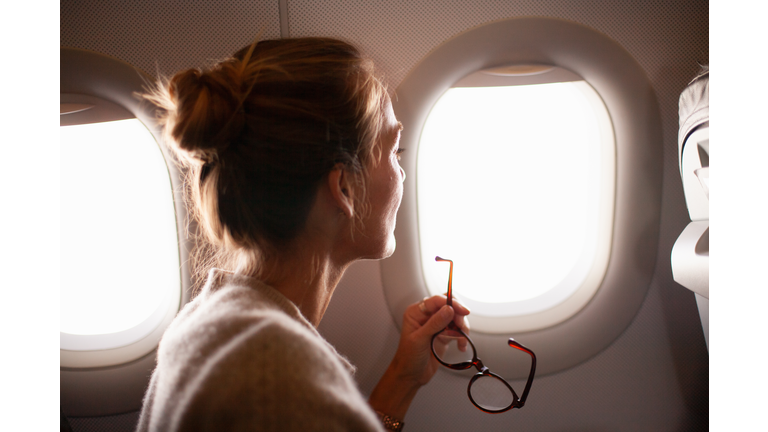 Woman looking through airplane window, holding reading glasses