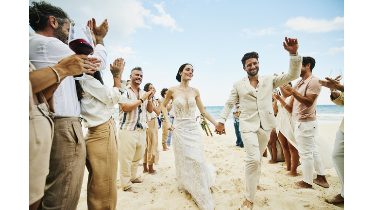 Wide shot of bride and groom walking down aisle after wedding ceremony on tropical beach while friends and family celebrate