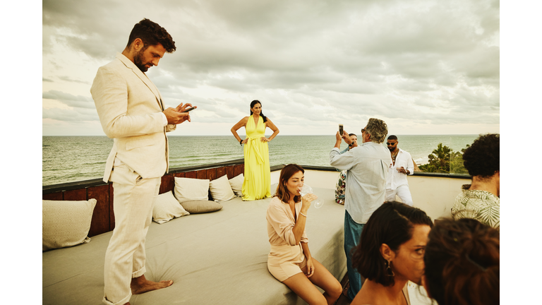 Wide shot of senior man taking photo of wife during sunset rooftop party after wedding at tropical resort