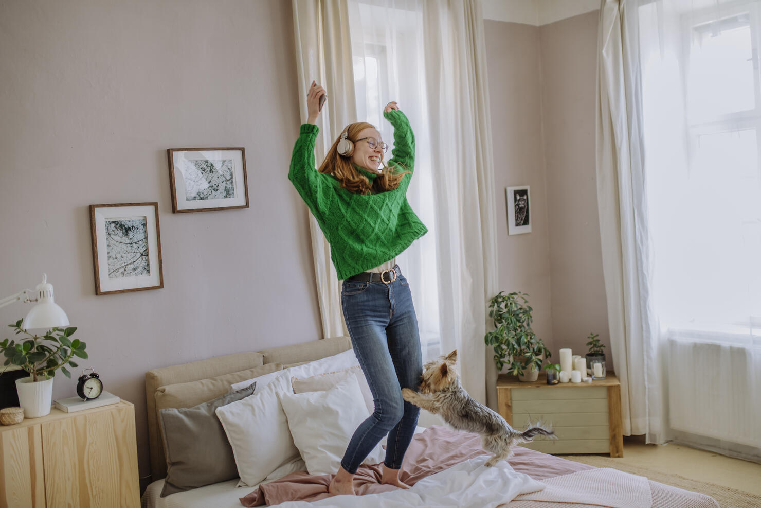 Happy woman listening music through wireless headphones dancing with arms raised by dog on bed at home