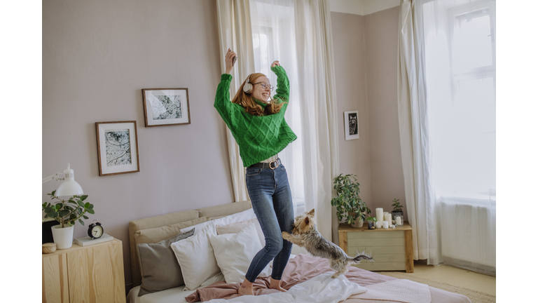 Happy woman listening music through wireless headphones dancing with arms raised by dog on bed at home