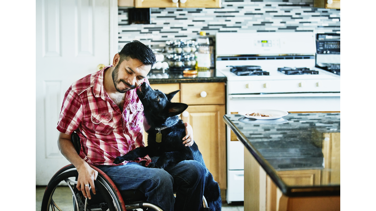 Smiling man in wheelchair having face licked by dog while hanging out in kitchen