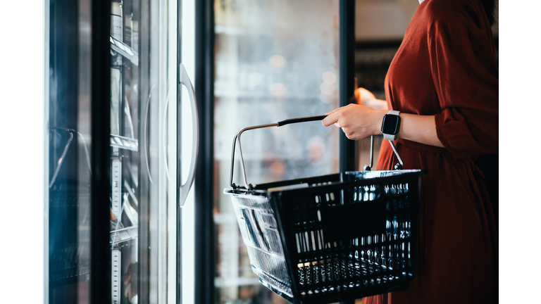 Mid-section of young Asian woman carrying a shopping basket, standing along the cold produce aisle, shopping for frozen food and daily necessities in supermarket. Healthy eating lifestyle. Making healthier food choices. Home cooking lifestyle