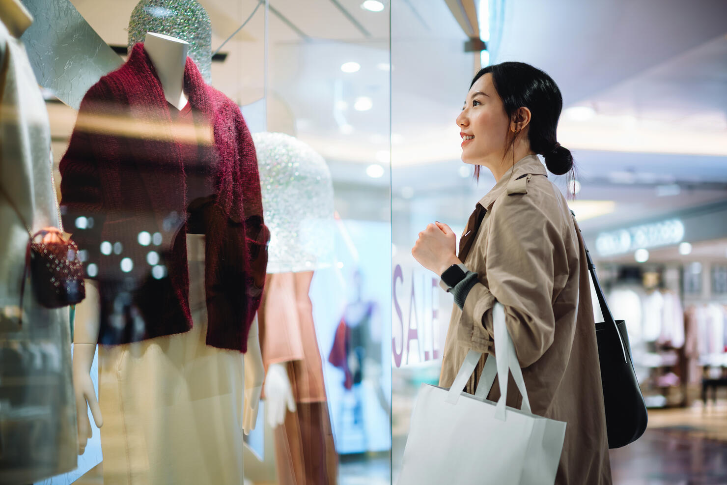 Beautiful smiling young Asian woman carrying a shopping bag, standing outside a boutique looking at window display in a shopping mall in city. Sale season, festive shopping atmosphere