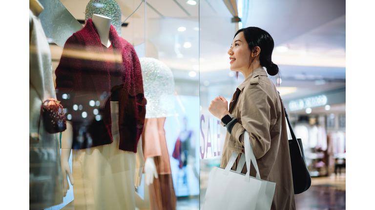 Beautiful smiling young Asian woman carrying a shopping bag, standing outside a boutique looking at window display in a shopping mall in city. Sale season, festive shopping atmosphere