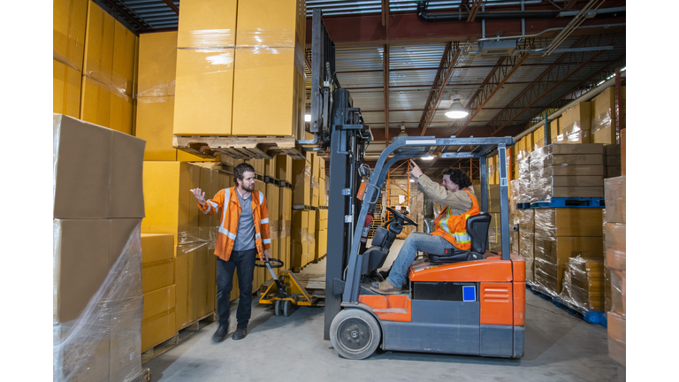 An industrial warehouse workplace safety topic.  A worker in the danger zone, walking under a raised forklift carrying a load.