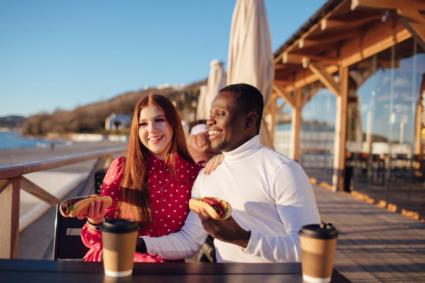 a young European woman with red hair and her black boyfriend are sitting in a cafe on an open terrace by the sea and eating a hot dog and coffee