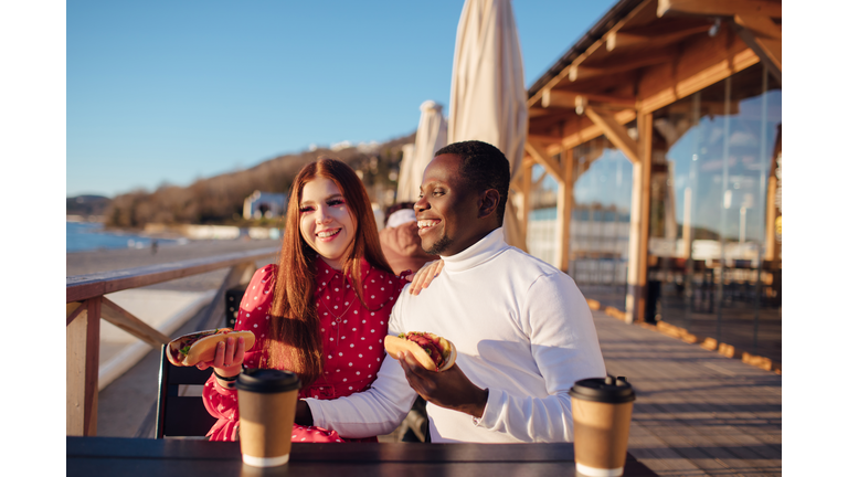 a young European woman with red hair and her black boyfriend are sitting in a cafe on an open terrace by the sea and eating a hot dog and coffee