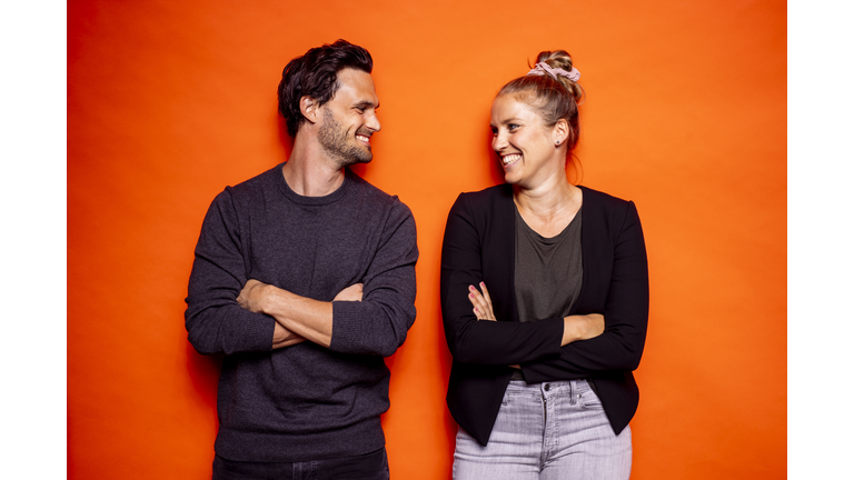 Happy mid adult couple looking at each other while standing with arms crossed against orange background