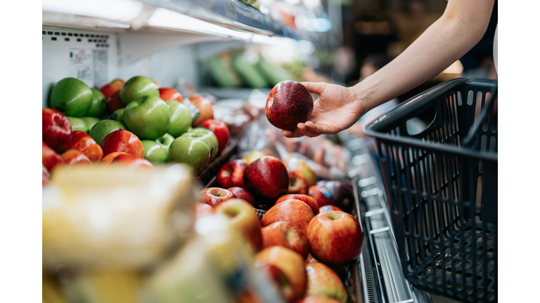 Cropped shot of young Asian woman choosing fresh organic fruits in supermarket. She is picking a red apple along the produce aisle. Routine grocery shopping. Healthy living and eating lifestyle