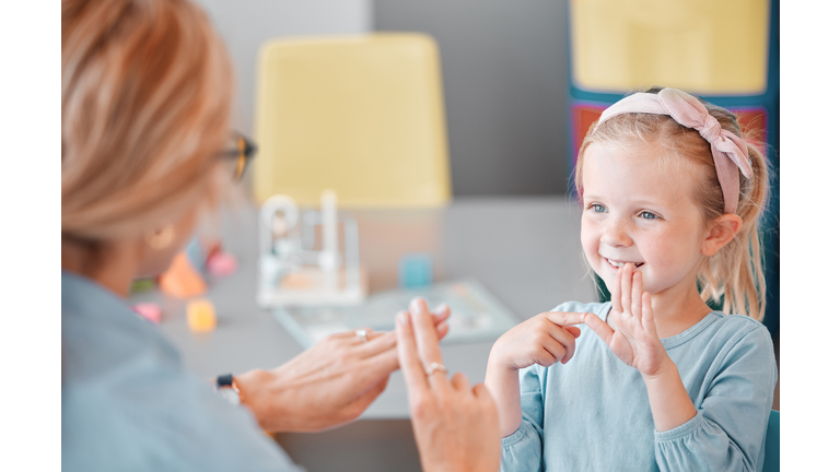 Adorable smiling little caucasian girl standing and using sign language to communicate with child psychologist in a clinic. Mental health professional teaching deaf kid to speak through hand gestures