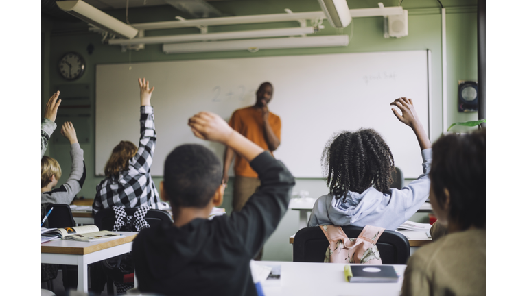 Rear view of multiracial students raising hands in classroom during lecture