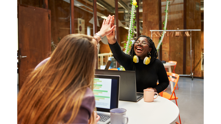 Two work colleagues giving each other high-five to express success while sitting at their workstations