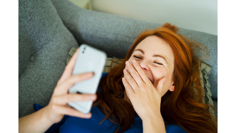 Woman lying on sofa using smart phone, smiling behind hand