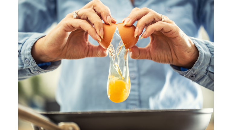 A detail of cracked egg falling into the pan as woman holds egg shells in both hands.