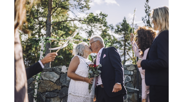 Senior bride with bouquet kissing groom on mouth amidst guest during wedding celebration