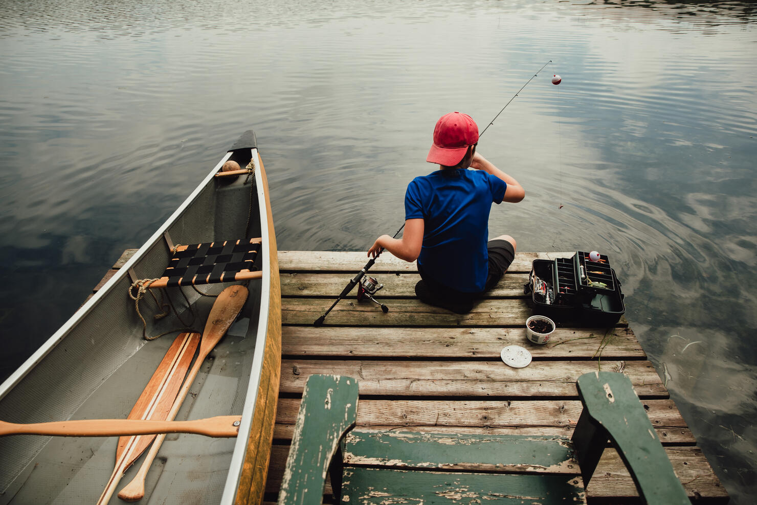 Teen boy fishing from a dock on a lake with tackle box in the summer.