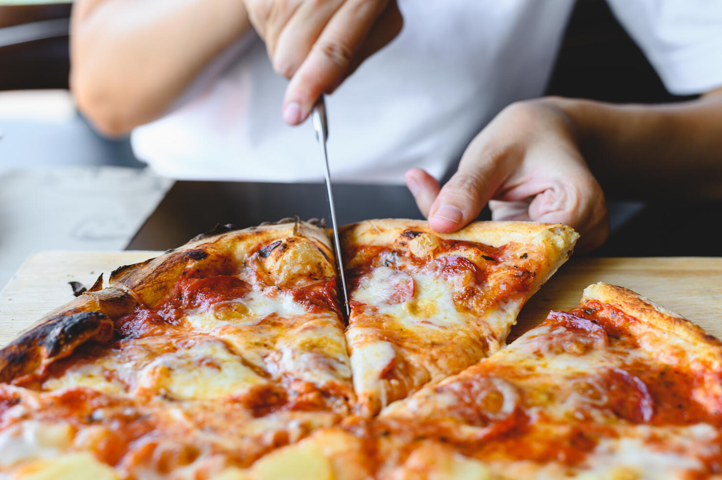 Wood fire cheesy pizza slice in hand. Traditional Hawaiian and Salami pizza savory dish flavour of origin Italian restaurant. Wood-Fired Oven Pizza with people holding and cutting knife background