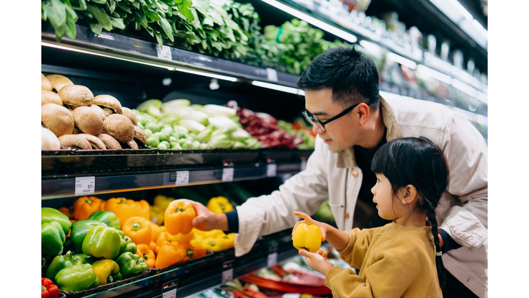 Young Asian father and her little daughter grocery shopping in supermarket. They are choosing fresh organic bell peppers together along the produce aisle. Fruits and vegetables shopping. Routine grocery shopping. Going green and healthy eating lifestyle