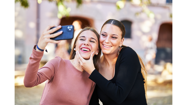 Two cheerful women smiling very affectionately looking at a phone while taking a selfie, front view