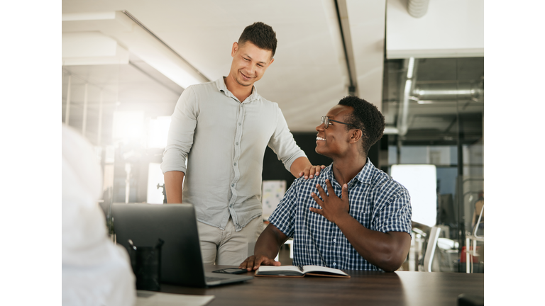 Two diverse businessmen talking at a desk discussing new project on laptop. Mentor complimenting colleague on a job well done and being a fast learner in modern office