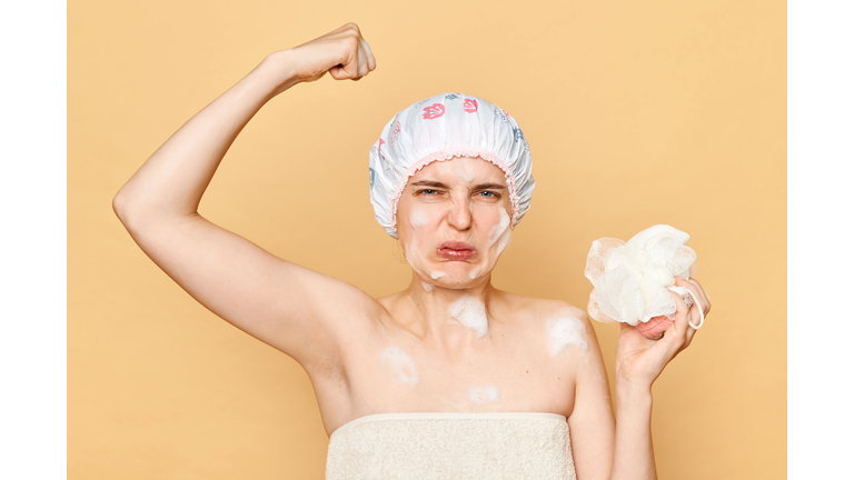 Delicate morning pampering. Displeased confused young woman in shower cap and wrapped in towel frowning face smelling armpit sweat isolated over beige background