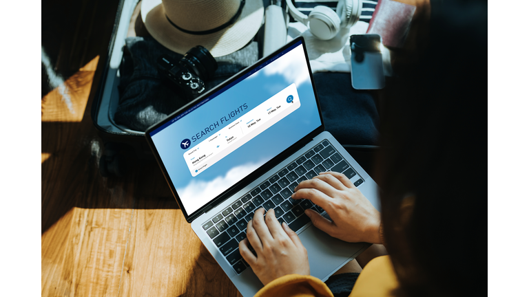 Young Asian woman shopping online for flight ticket on airline website with laptop at home, sitting next to suitcase with sun hat, camera, clothings, headphones, smartphone and passport. Getting ready for vacation. Travel and vacation concept