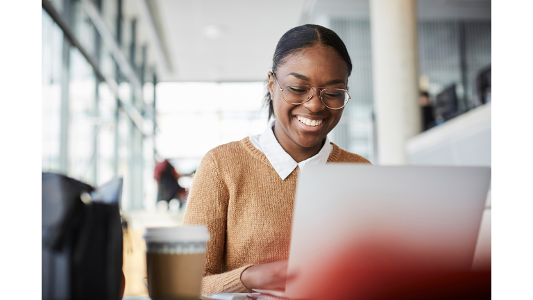 Smiling young female student using laptop while studying at table in university cafeteria