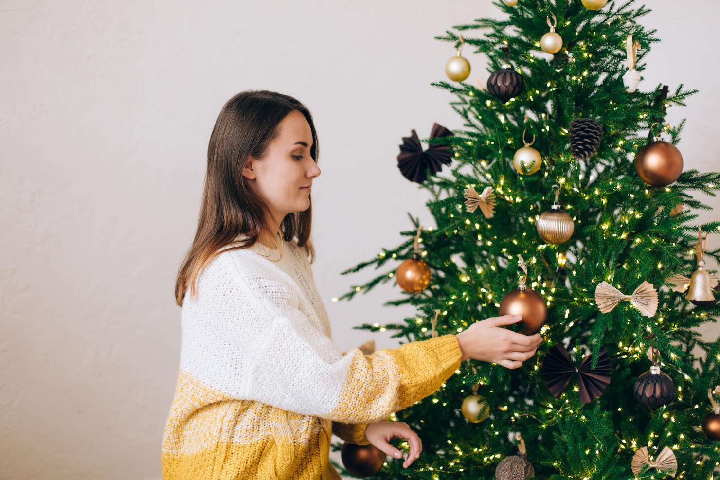 Smiling woman in white-yellow knitted sweater is decorating green Christmas tree with bronze baubles against white wall. New Year celebration concept. Front view