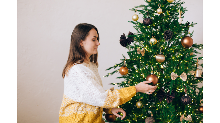 Smiling woman in white-yellow knitted sweater is decorating green Christmas tree with bronze baubles against white wall. New Year celebration concept. Front view